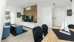 A modern living room with a flat-screen TV, a wooden panel wall, a dining area with a sailboat model, and a small plant. - Serenity - Wave Resort & Spa (Międzyzdroje)