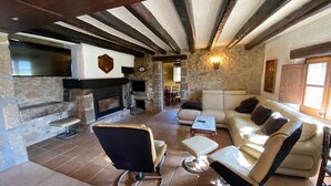 Une cheminée en pierre, un canapé et un téléviseur dans un salon chaleureux aux poutres apparentes.
