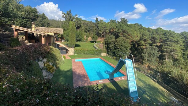 Une piscine avec un toboggan, entourée de verdure, et une maison avec un toit en tuiles.