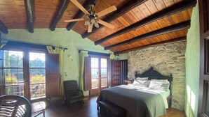Une chambre à coucher avec un lit, une chaise, un ventilateur de plafond en bois et un mur en pierre.