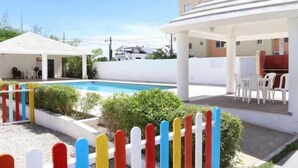 Zona de piscina amb una tanca blanca, zona d'seients coberta i un edifici amb teulada plana.