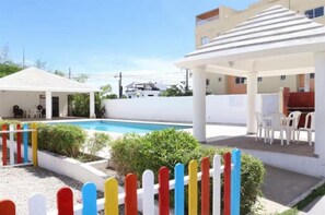 A pool area with a white fence, a covered seating area, and a building with a flat roof. - Palmarés de caleta (La Romana)
