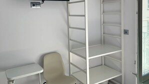 A room with a TV mounted on the wall, a white desk, a chair, and a white shelving unit with a wooden hanger. - Hostal Palacio Leganitos (Madrid)