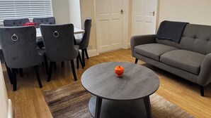 A living room with a grey sofa, a round coffee table with an orange object, a dining table with chairs, and a white door. - Cosy 3 bedroom house in Sale, Manchester (Sale)