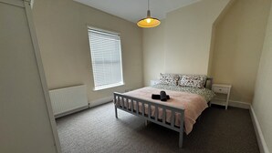 A bedroom with a bed, a bedside table, a nightstand, a radiator, a window with blinds, and a hanging light fixture. - Cosy 3 bedroom house in Sale, Manchester (Sale)
