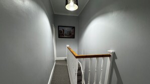 A narrow hallway with a wooden handrail and white balusters, carpeted floor, and a light fixture on the ceiling. - Cosy 3 bedroom house in Sale, Manchester (Sale)