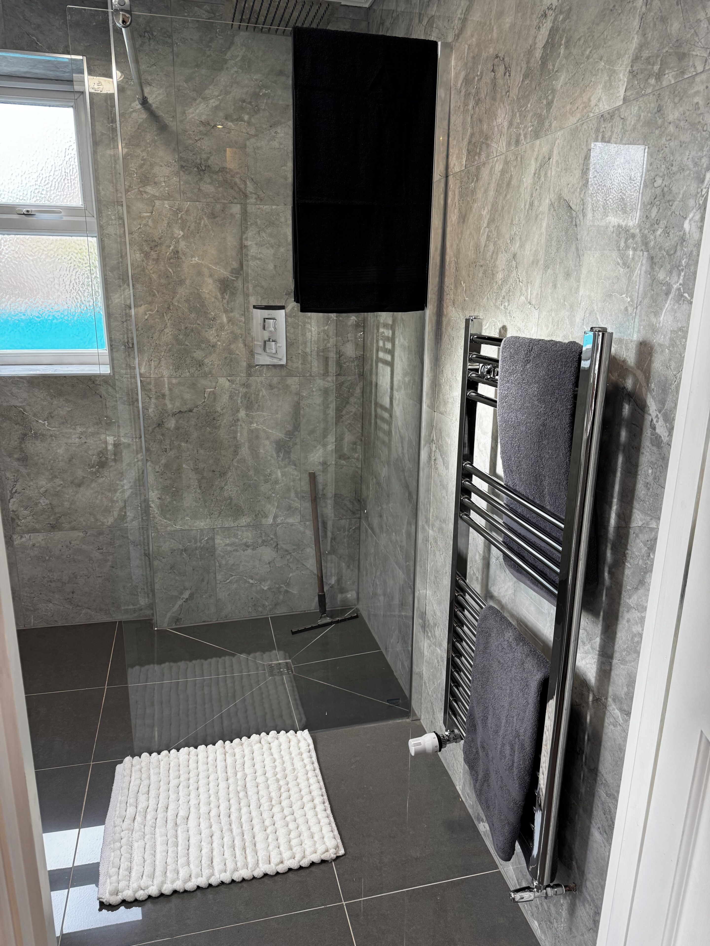 A bathroom with a shower, a towel rack, a window, and a white rug.