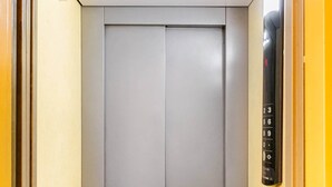 Een lift met een bedieningspaneel aan de rechterkant, een gesloten liftdeur en een ventilatie rooster onderaan.