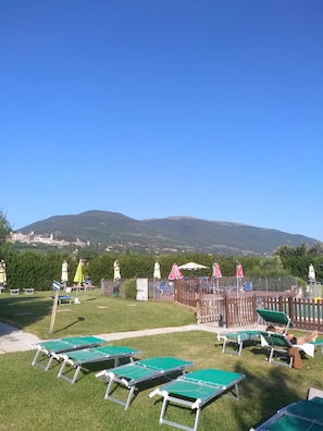 Un espacio al aire libre con césped, tumbonas y sombrillas, una valla de madera y una vista de la montaña al fondo.