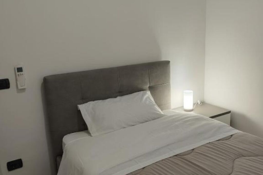 A neatly made bed with a grey headboard, a bedside table with a lamp, and a wall-mounted thermostat.