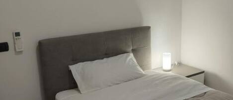 A neatly made bed with a grey headboard, a bedside table with a lamp, and a wall-mounted thermostat.