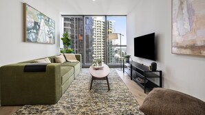 A modern living room with a sofa, a coffee table, a TV, and a large window with a city view. - Skyline & Sea Luxe Heated Pool, Gym, Sauna, Steam (Melbourne)