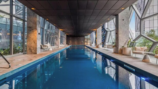 A long indoor swimming pool with clear glass walls, modern design, and seating areas. - Skyline & Sea Luxe Heated Pool, Gym, Sauna, Steam (Melbourne)