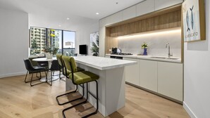 A modern kitchen with a central island, dining area with chairs, and a view of buildings outside. - Skyline & Sea Luxe Heated Pool, Gym, Sauna, Steam (Melbourne)