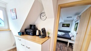 Een compact keukenhoekje met een magnetron, koffiezetapparaat en waterkoker, een nachtkastje met een plant en een bed met uitzicht op een bos.