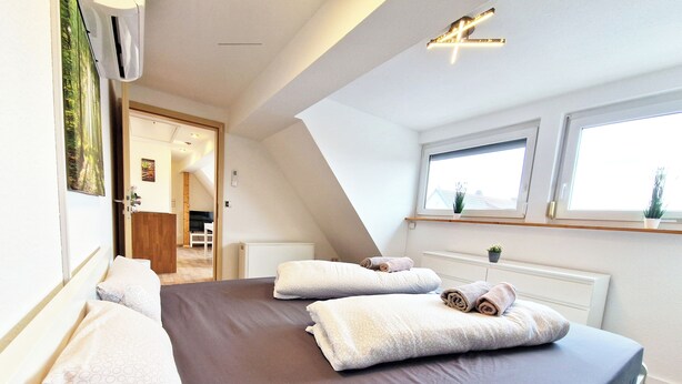 Een slaapkamer met een groot bed, een raam, een radiator en een deur naar een andere kamer.
