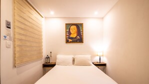 Habitación de hotel con cama, mesita de noche con lámpara, un cuadro en la pared y ventana con persianas.