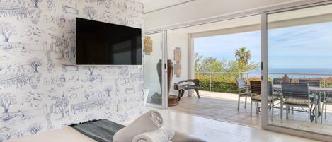 A hotel room with a bed, a flat-screen TV, a balcony with a table and chairs, and a view of the sea.