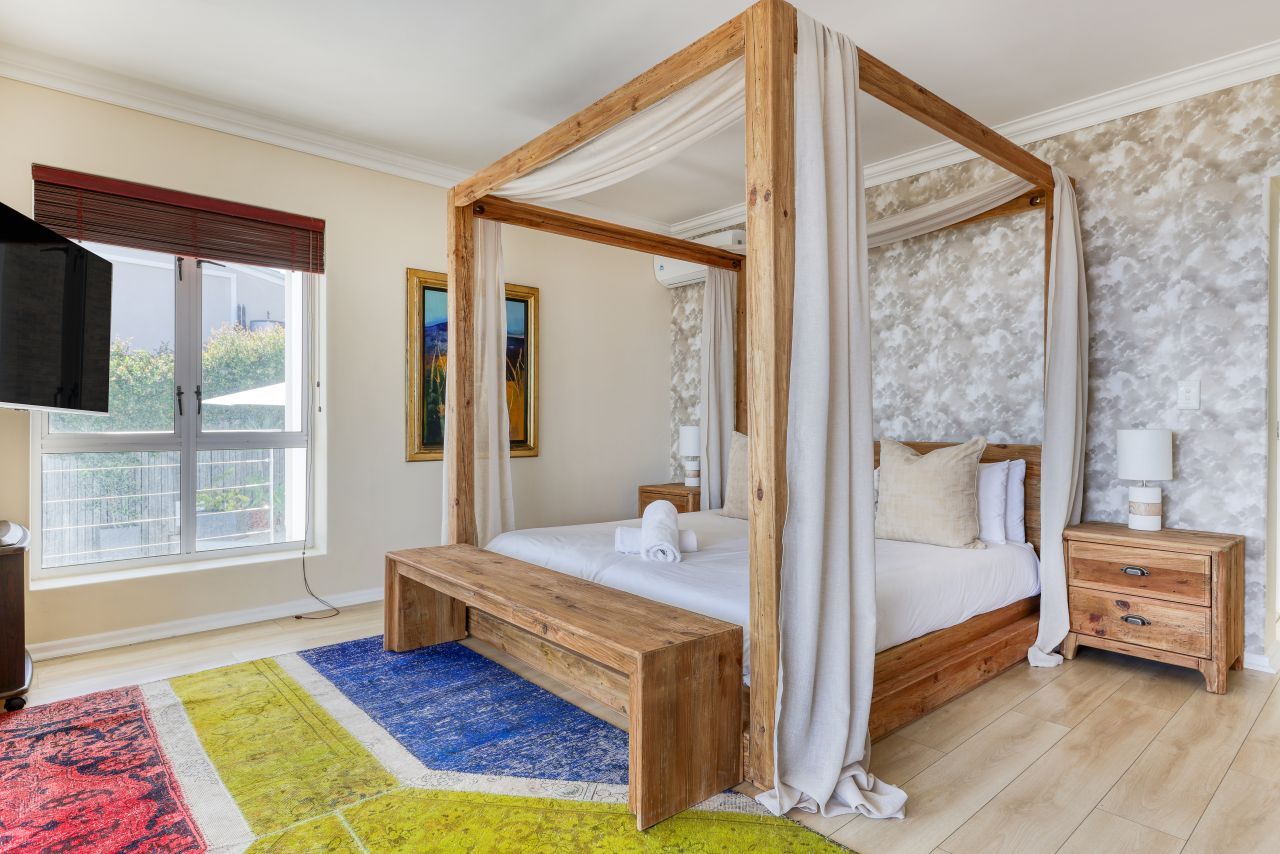 A four-poster bed with a wooden frame and white bedding, a bedside table with a lamp, a bench at the foot of the bed, and a colorful rug.