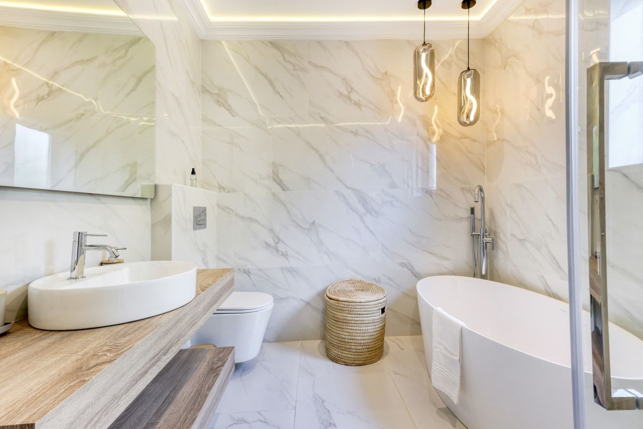 Ein modernes Badezimmer mit Marmorwand, Waschtisch aus Holz, einem weißen Waschbecken, einer Toilette und einer freistehenden Badewanne.