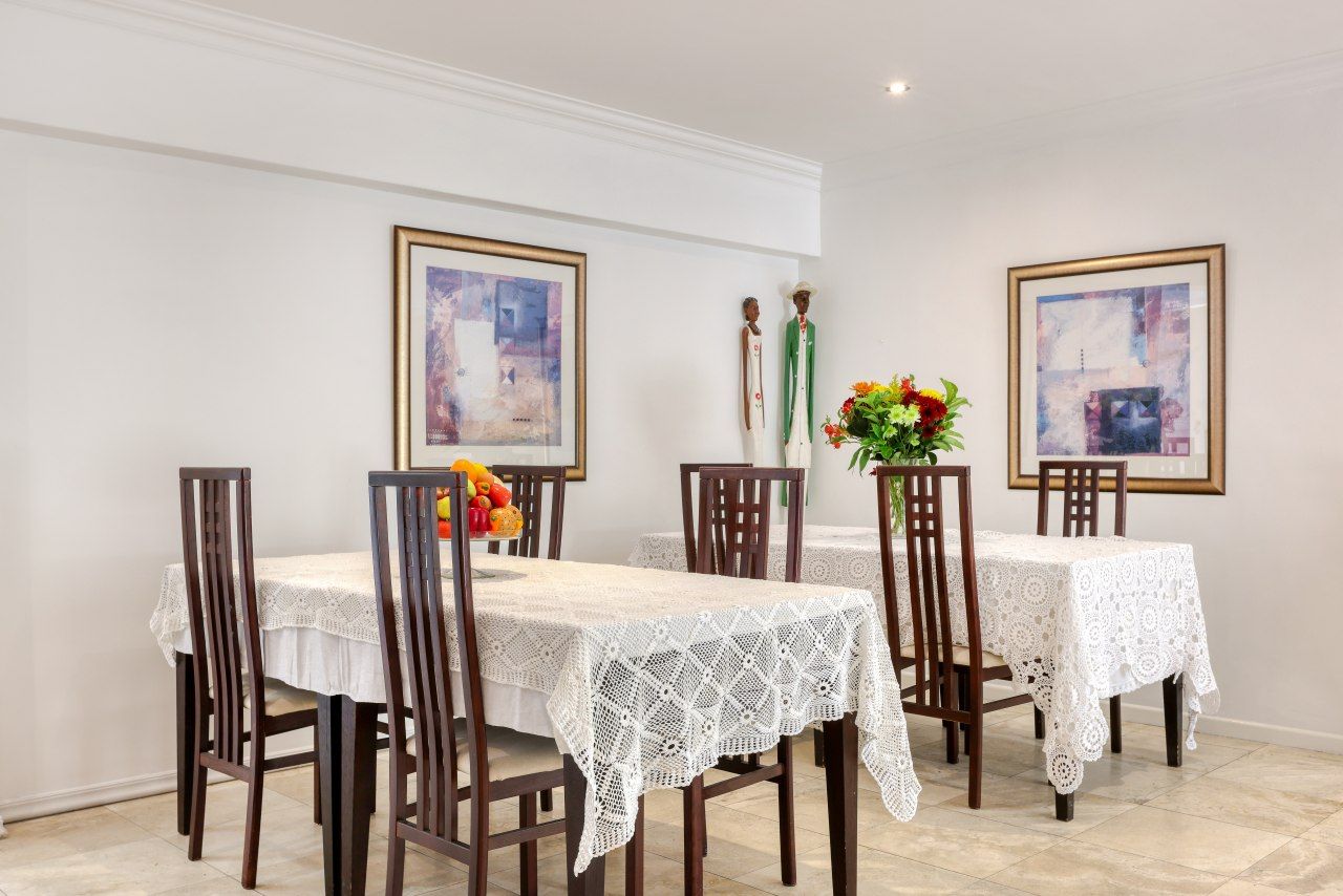 Un coin repas avec une table dressée pour quatre personnes, agrémenté d’une nappe en dentelle blanche, de chaises en bois et d’œuvres d’art encadrées accrochées aux murs.