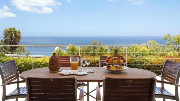 Een ronde tafel met stoelen, een fruitschaal, een glas sinaasappelsap en een keramieken kan op een balkon met uitzicht op zee.