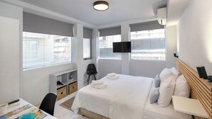 A hotel room with a bed, a desk with a chair, a TV, and a window with blinds. - BES 6 - Minimal Studio by GHH (Kesariani)