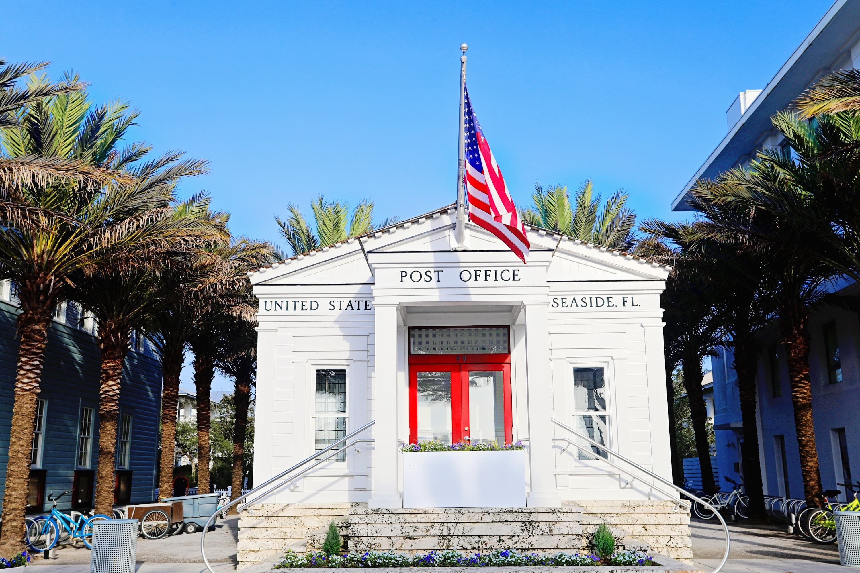 A white post office with a red door, an American flag, and palm trees in the background.