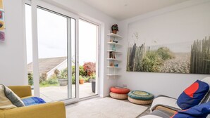 A modern living room with a sliding glass door leading to a garden, a sofa, a chair, and a bookshelf with decorative items. - Wootton Gray (Penzance)