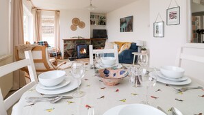 A dining area with a table set for four, a fireplace, a bookshelf, and a sofa in the background. - Wootton Gray (Penzance)