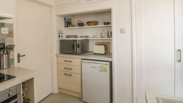 A compact kitchen with a microwave, toaster, and refrigerator, along with a set of drawers and a shelf above. - Wootton Gray (Penzance)