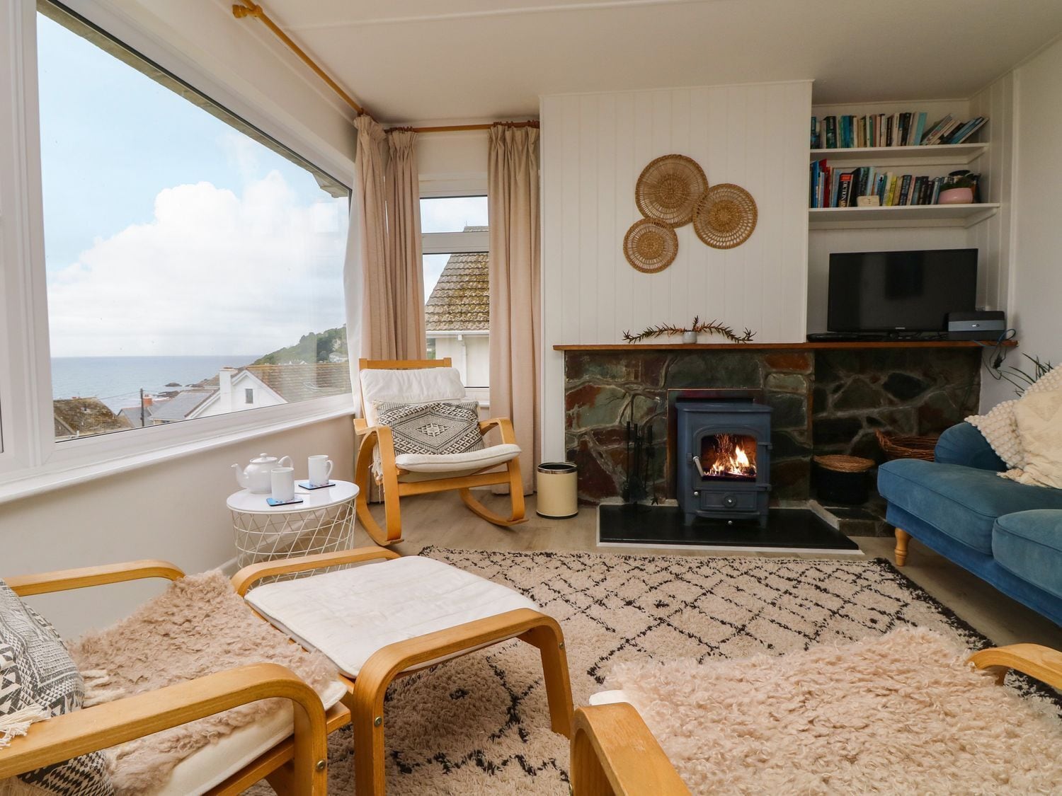 A cozy living room with a stone fireplace, a blue sofa, a wooden rocking chair, and a view of the sea.