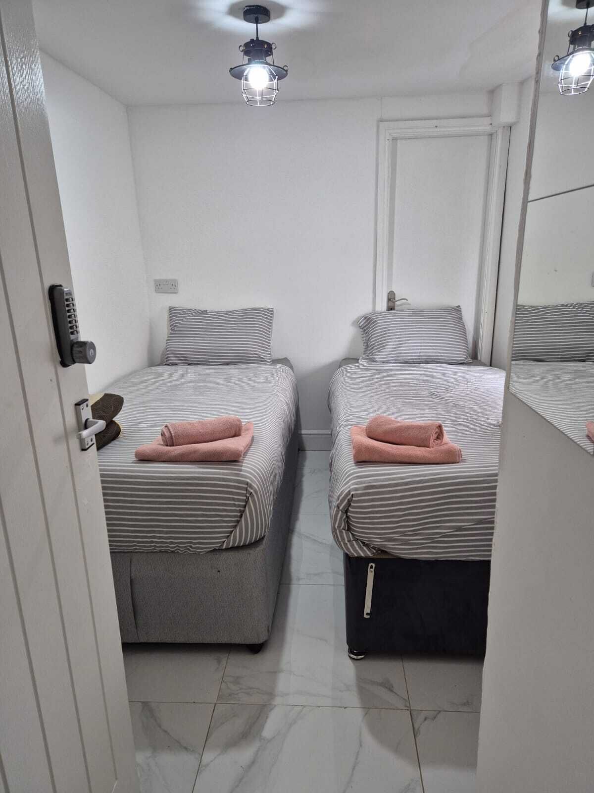 A room with two beds, each with striped bedding and a pink towel, a door with a handle, and a ceiling light fixture.