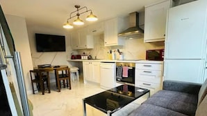 A modern kitchen with white cabinets, a dining area with a table and chairs, a gray sofa, and a TV mounted on the wall. - Private room in Little Bridgeford (Stafford)