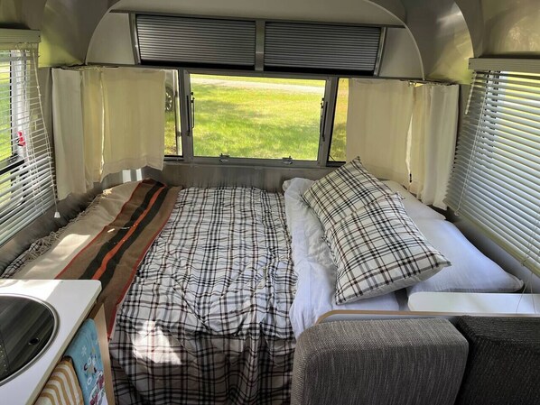 Een compact RV-interieur met een bed, een kleine keuken en een raam met uitzicht op een grasveld.