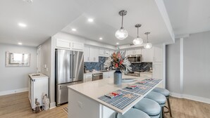 Una cocina moderna con electrodomésticos de acero inoxidable, una isla central con taburetes de bar, iluminación colgante y un jarrón decorativo.