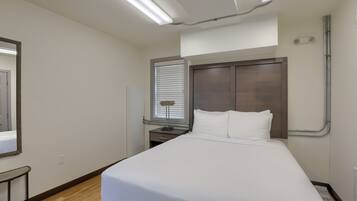 A hotel room with a bed, a nightstand, a lamp, a window with blinds, and a mirror on the wall.