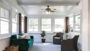 Une véranda avec un ventilateur de plafond, de grandes fenêtres, un canapé vert, un fauteuil en osier et un tapis à motifs.