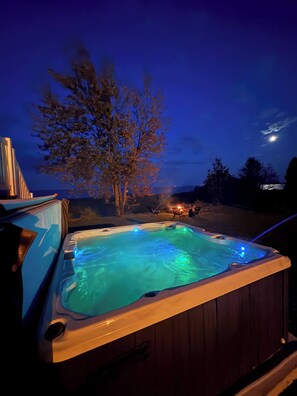 A hot tub with a wooden exterior and a pool of water, surrounded by trees and a clear sky.