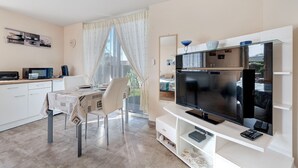 A modern living room with a flat-screen TV, a dining table with chairs, a kitchen area with appliances, and a large window with curtains.
