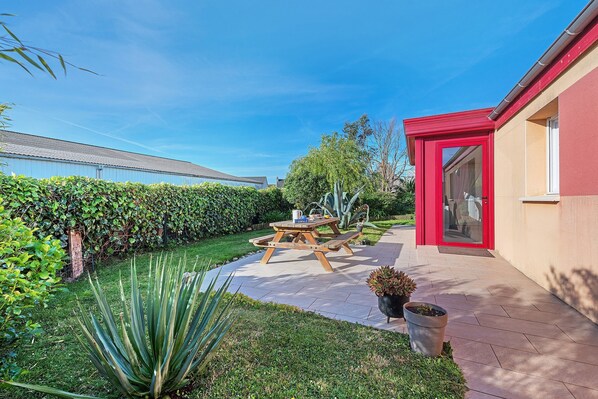 A patio with a wooden table and benches, a red door, and a well-maintained garden.