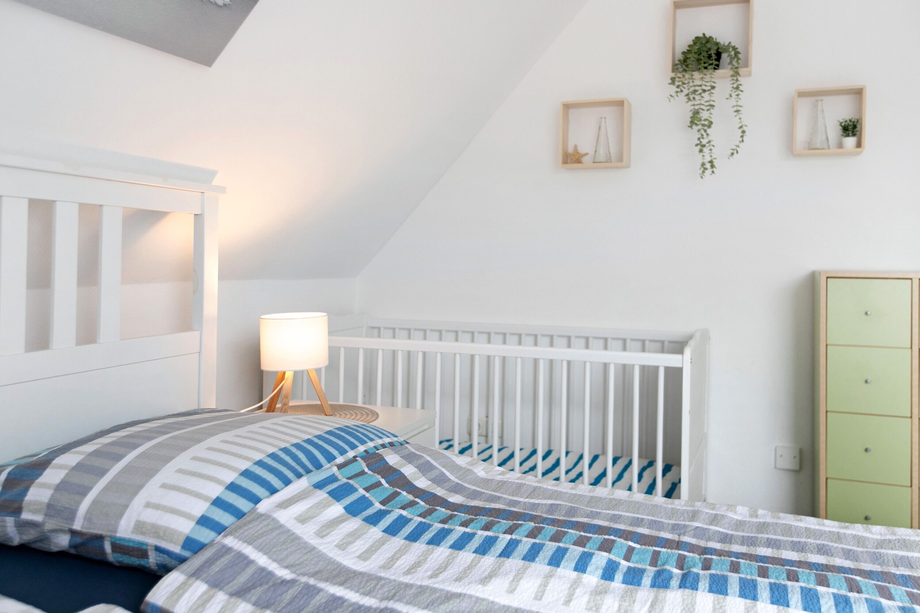 Una habitación con una cuna, una cama con ropa de cama a rayas, una lámpara de noche y un armario verde.