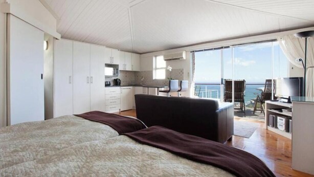 Ein Schlafzimmer mit einem großen Bett, einem dunklen Sofa, einer Küchenzeile und einem Balkon mit Meerblick.
