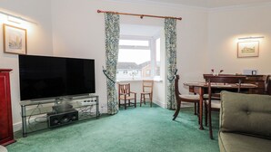 A living room with a flat-screen TV, a glass TV stand, a dining table with chairs, and a green carpet. - The Flat House (Taunton)