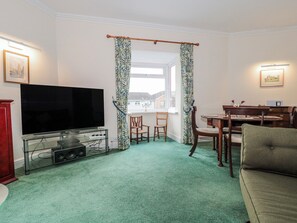 A living room with a flat-screen TV, a glass TV stand, a dining table with chairs, and a green carpet. - The Flat House (Taunton)