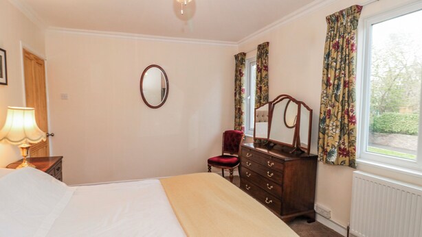 A bedroom with a bed, a dresser with a mirror, a chair, a lamp, and a window with curtains. - The Flat House (Taunton)