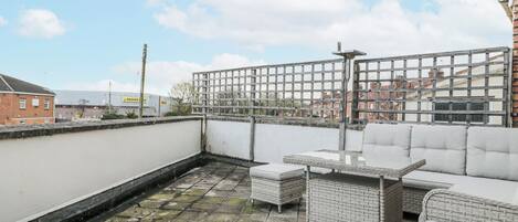A rooftop terrace with a white wall, a metal fence, and a seating area with wicker furniture.