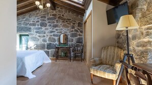 A room with a stone wall, wooden ceiling beams, a bed, a desk with a chair, a lamp, and a television. - CDC Casa do Capitán Leiro: Relax Among Vineyards and Forests in Ribeiro (Cubilledo)