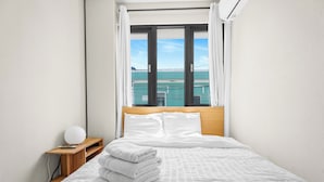 Un letto rifatto con lenzuola e cuscini bianchi, un comodino con una lampada e una finestra con tende.