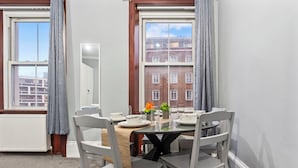 Uma área de refeições com uma mesa posta para quatro pessoas, cadeiras brancas e vista para um edifício de tijolos através de grandes janelas.
